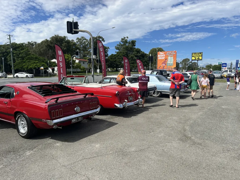 Red classic fastback and convertible leading a row of classic cars, with visitors browsing at a car meet