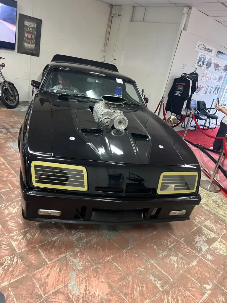 Black Mad Max Interceptor replica behind a red rope on the museum floor, with T-shirts on display behind